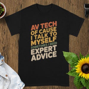 May include: Black t-shirt with the text "AV TECH OF CAUSE I TALK TO MYSELF SOMETIMES I NEED EXPERT ADVICE" in orange, yellow, and white. The shirt is on a wooden surface with a small plant and a sunflower.
