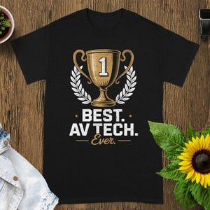 May include: Black t-shirt with a gold trophy graphic and the text "BEST. AV TECH. Ever." The trophy has the number "1" on it. The design is surrounded by a white laurel wreath. A sunflower and a small plant are in the background.