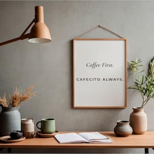 Puede incluir: Un escritorio de madera con un cartel enmarcado que dice "Coffee First. Cafecito Always." En el escritorio hay varias tazas de cerámica, jarrones y un libro abierto. Una lámpara de madera y plantas completan la escena.