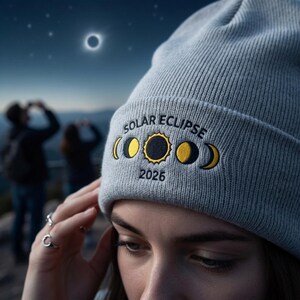 May include: A light gray beanie with the words "SOLAR ECLIPSE" and the year "2026" embroidered in yellow and black. The design includes phases of the moon. The background shows a solar eclipse and people looking up.