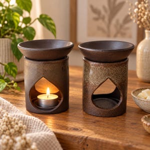 May include: Two cylindrical ceramic oil burners with dark brown, speckled glaze finishes. Each burner features a teardrop-shaped cutout for a tea light candle. One burner contains a lit tea light candle, while the other is empty.