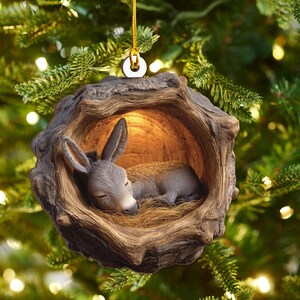 May include: A Christmas ornament featuring a sleeping donkey nestled inside a wooden hollow. The donkey is gray and brown, resting on a bed of straw. The ornament is suspended by a gold string.
