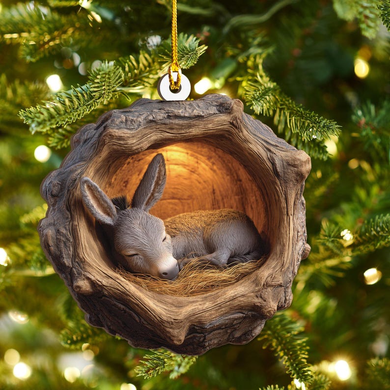 May include: A Christmas ornament featuring a sleeping donkey nestled inside a wooden hollow. The donkey is gray and brown, resting on a bed of straw. The ornament is suspended by a gold string.