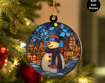 Puede incluir: Adorno navideño redondo con un diseño de muñeco de nieve. El muñeco de nieve lleva un sombrero de copa azul, una bufanda a rayas y una nariz de zanahoria. El fondo representa una escena de pueblo. El adorno está impreso por ambas caras.