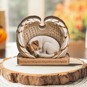 Op de afbeelding: Een decoratief houten herdenkingsornament met een slapende Jack Russell Terrier opgerold in een paar grote, gedetailleerde engelvleugels. De basis is gegraveerd met de tekst: "You may have left my arms, but you'll never leave my heart."