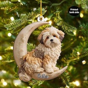 May include: A Christmas tree ornament depicting a fluffy dog on a crescent moon. The dog has brown, white, and tan fur. The ornament is inscribed with the name "Bella" and the text "Printed both side".