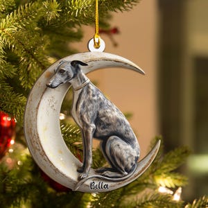 Puede incluir: Adorno navideño con un galgo sentado en una luna creciente. El perro tiene un pelaje detallado y lleva un collar. El adorno está suspendido por una cuerda dorada y tiene el nombre "Bella".
