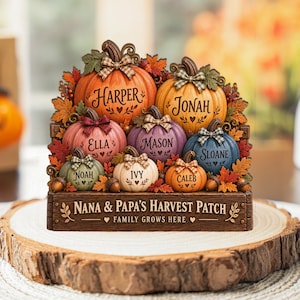 Puede incluir: Un adorno de mesa decorativo con siete calabazas coloridas de distintos tamaños, cada una grabada con un nombre. Las calabazas están decoradas con lazos de cuadros y rodeadas de hojas de otoño y bellotas. La base dice NANA & PAPA'S HARVEST PATCH.