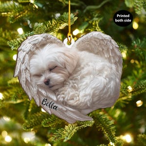 Op de afbeelding: Een hartvormige kerstversiering met een slapende witte hond in engelen vleugels. De versiering hangt aan een gouden draad en heeft de naam "Bella" erop gedrukt. De tekst "Printed both side" is ook zichtbaar.