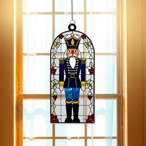 May include: A stained glass window hanging ornament featuring a nutcracker in a blue and red uniform with a white beard and mustache. The ornament is surrounded by a floral design with red and white flowers.
