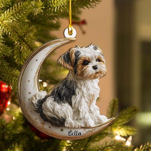 May include: A dog-themed Christmas ornament. The ornament features a small, fluffy dog sitting on a crescent moon. The moon is white with gold trim and small star accents. The dog has brown, black, and white fur. The name "Bella" is written on the moon.