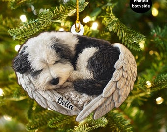 May include: A Christmas ornament featuring a sleeping dog nestled in angel wings. The dog has black and white fur, and the wings are a light beige color. The name "Bella" is printed on the wings. The ornament is hanging from a gold string.