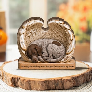 Può includere: Una targa decorativa in legno raffigurante un cane maculato marrone e bianco raggomitolato all'interno di un paio di grandi ali d'angelo dettagliate. La base è incisa con il testo, You may have left my arms, but you'll never leave my heart, e Bella.