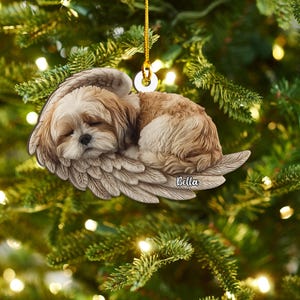May include: A decorative Christmas ornament featuring a sleeping dog with angel wings. The dog has brown and white fur and is resting on detailed, feathery wings. The name "Bella" is written on the wing. The ornament hangs from a gold string.
