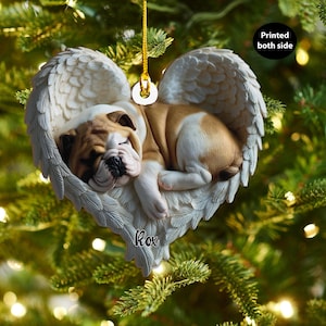 May include: A heart-shaped Christmas ornament featuring a sleeping bulldog puppy nestled in angel wings. The ornament is white and cream with a gold hanging cord. The puppy is brown and white. The text "Printed both side" is visible.