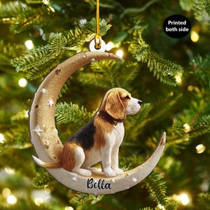Op de afbeelding: Een kerstboomornament met een beagle hond die op een gouden halve maan zit, versierd met witte sterren. De hond heeft bruin, zwart en wit haar. De naam "Bella" staat op de maan.