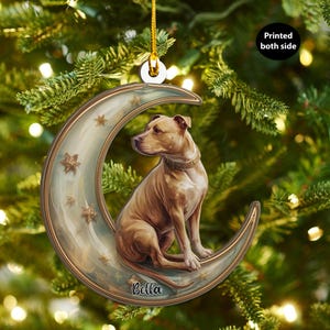 Może przedstawiać: Ozdoba świąteczna przedstawiająca brązowego psa siedzącego na półksiężycu ze złotymi gwiazdami. Ozdoba ma złote obramowanie i złoty sznurek do zawieszenia. Na księżycu widnieje napis "Bella". Widoczny jest napis "Printed both side".