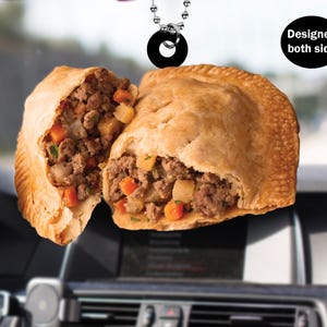 May include: A car air freshener shaped like a savory meat pie with a flaky crust. The pie is cut in half, revealing a filling of ground meat, vegetables, and gravy. The air freshener is designed to hang from the rearview mirror. "Designed both side" is printed on the white background.