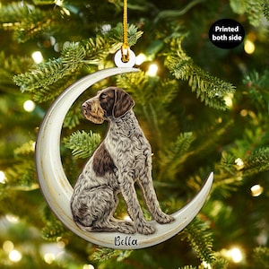 May include: A crescent moon-shaped dog ornament. The ornament depicts a brown and white dog illustration. The name "Bella" is written on the moon. The ornament hangs from a gold string. The text "Printed both side" is visible on the ornament.