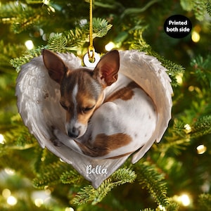 Può includere: Un ornamento natalizio con un cagnolino in un design ad ali d'angelo a forma di cuore. Il cane ha il pelo marrone e bianco. L'ornamento è sospeso da un filo d'oro e ha il nome "Bella". L'ornamento è stampato su entrambi i lati.