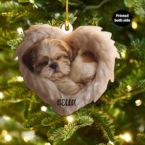 Può includere: Un ornamento natalizio a forma di cuore con un cucciolo di Shih Tzu addormentato in ali d'angelo. Il cucciolo ha il pelo marrone e bianco e l'ornamento è personalizzato con il nome "BELLA". È appeso a un filo d'oro.