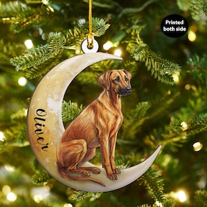 May include: A crescent moon-shaped Christmas ornament featuring a brown dog with a black nose and a name written in cursive. The ornament is painted with a light beige color and has a gold string for hanging. The text "Printed both side" is also visible.
