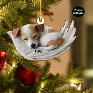 Puede incluir: Un adorno navideño con un cachorro durmiendo con pelaje marrón y blanco, descansando sobre un diseño de ala de ángel blanca. El adorno tiene el nombre "LUNA" impreso y cuelga de una cuerda dorada.