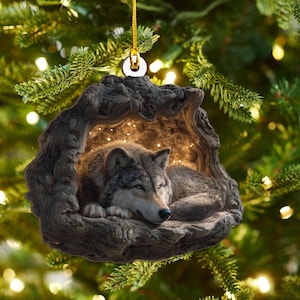 May include: A detailed Christmas ornament featuring a resting wolf inside a carved wooden cave. The wolf has gray and brown fur, and the cave is a dark brown color. The ornament hangs from a gold string.
