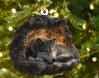 May include: A detailed Christmas ornament featuring a resting wolf inside a carved wooden cave. The wolf has gray and brown fur, and the cave is a dark brown color. The ornament hangs from a gold string.