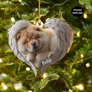 Op de afbeelding: Een hartvormige kerstversiering met een slapende Chow Chow-hond genesteld in engelen vleugels. De versiering is lichtbruin en wit, met de naam "Bella" erop gedrukt. De tekst "Printed both side" is ook zichtbaar.