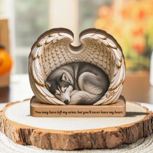 May include: A layered wooden memorial ornament featuring a sleeping husky dog nestled within a pair of carved angel wings. The base is engraved with the text, "You may have left my arms, but you'll never leave my heart," and the name "Bella."