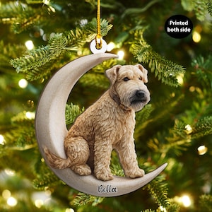 May include: A beige dog ornament sitting on a crescent moon. The dog has a brown collar and is sitting on a beige crescent moon. The name "Bella" is printed on the moon. The ornament is hanging from a gold string. The text "Printed both side" is on the ornament.
