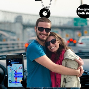 Puede incluir: Una foto en blanco y negro de una pareja abrazándose en un coche. El hombre lleva gafas de sol y una camisa azul. La mujer lleva gafas de sol y una chaqueta beige sobre una bufanda rosa. La foto está impresa en un charm de coche redondo y negro con una cadena plateada.