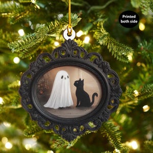 Peut inclure: Ornement d'Halloween encadré en noir avec une illustration de fantôme et de chat noir. L'ornement est suspendu à une ficelle dorée sur fond de branches de sapin de Noël vertes. Le texte "Printed both side" est visible.