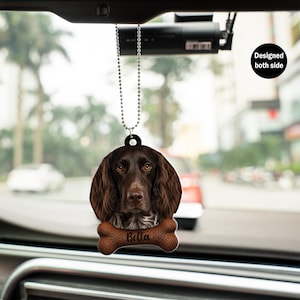 May include: A car hanging ornament featuring a brown dog with long, wavy fur and a bone-shaped tag. The tag is inscribed with the name "Bella." The ornament is designed to be visible from both sides and hangs from a silver beaded chain.