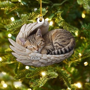 May include: A Christmas tree ornament depicting a sleeping tabby cat cradled in angel wings. The beige ornament has the name "Bella" on it. It hangs from a gold string. The ornament is surrounded by green fir tree branches.