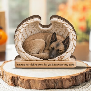 May include: A memorial plaque featuring a tan dog curled up inside a pair of white angel wings. The name Bella is engraved on the wing, and the base displays the text: You may have left my arms, but you'll never leave my heart.
