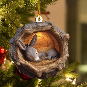 May include: A Christmas tree ornament featuring a sleeping donkey nestled inside a wooden hollow. The donkey is gray with closed eyes, resting on a bed of straw. The ornament is suspended by a gold string.