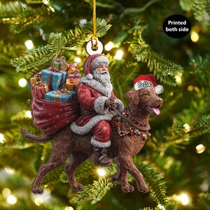 May include: A Christmas ornament featuring Santa Claus riding a brown dog with a Santa hat. Santa is carrying a sack of gifts. The dog has a name tag that says "Bella". The ornament is hanging from a gold string.