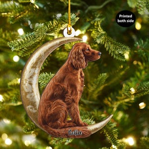 Op de afbeelding: Een kerstboomversiering met een bruine hond die op een gouden halve maan zit. De versiering heeft een gouden draad om op te hangen en de tekst "Bella" staat op de maan. De tekst "Printed both side" is ook zichtbaar.