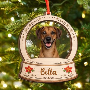 May include: A wooden Christmas ornament shaped like a snow globe featuring a brown dog with a happy expression. The base of the ornament is engraved with the name Bella and decorated with red poinsettia flowers. A red ribbon is attached to the top.