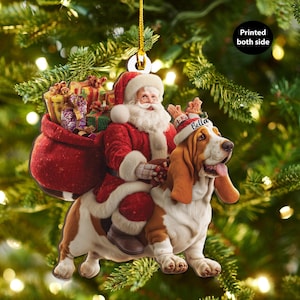 May include: A festive Christmas tree decoration depicting Santa Claus on a Basset Hound. Santa is in a red suit with a sack of presents. The dog wears a hat with "Bella" on it. The ornament hangs from a gold string.