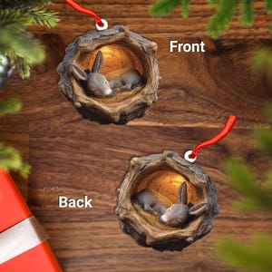 May include: Two Christmas ornaments featuring a sleeping donkey inside a rustic, hollowed-out log. The donkey is gray and brown, nestled on a bed of straw. Each ornament has a red ribbon for hanging.