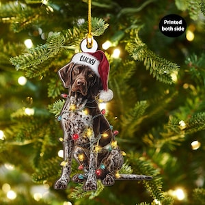 May include: A Christmas tree decoration depicting a brown and white dog wearing a Santa hat with the word "Lucky" and wrapped in colourful Christmas lights. The ornament is designed to be printed on both sides and is suspended from a gold string.