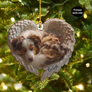 Può includere: Un ornamento natalizio a forma di cuore con un cane addormentato in ali d'angelo. Il cane ha il pelo marrone e bianco. L'ornamento ha un cordino dorato per appenderlo e il nome "Bella" è scritto in basso.