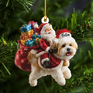 May include: A Christmas ornament featuring Santa Claus riding a fluffy dog named Bella. Santa is dressed in a red suit and carrying a sack of gifts. The dog is wearing a Santa hat and cape. The ornament hangs from a gold string.