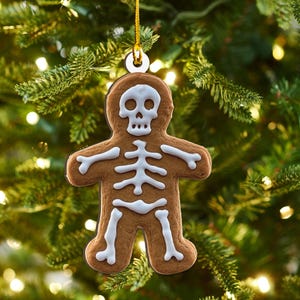 May include: A gingerbread man ornament shaped like a skeleton, with white icing details. The ornament is hanging from a gold string against a blurred background of a Christmas tree with lights.
