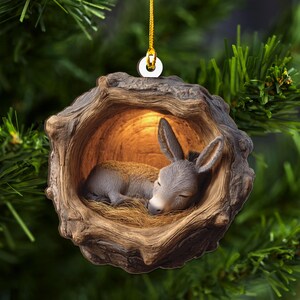 May include: A Christmas ornament featuring a sleeping donkey nestled in a wooden hollow. The donkey is gray with long ears, lying on straw. The ornament is brown with a gold hanging loop, set against a green background.