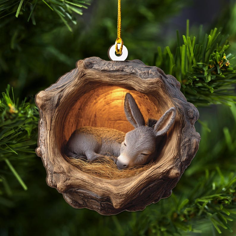 May include: A Christmas ornament featuring a sleeping donkey nestled in a wooden hollow. The donkey is gray with long ears, lying on straw. The ornament is brown with a gold hanging loop, set against a green background.