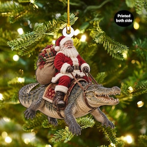 Könnte beinhalten: Ein Weihnachtsbaumschmuck, der den Weihnachtsmann mit weißem Bart und rotem Anzug auf einem grünen Alligator reitend zeigt. Der Weihnachtsmann trägt einen Jutesack voller Geschenke auf dem Rücken. Der Schmuck hat eine goldene Schnur und den Text Printed both side.